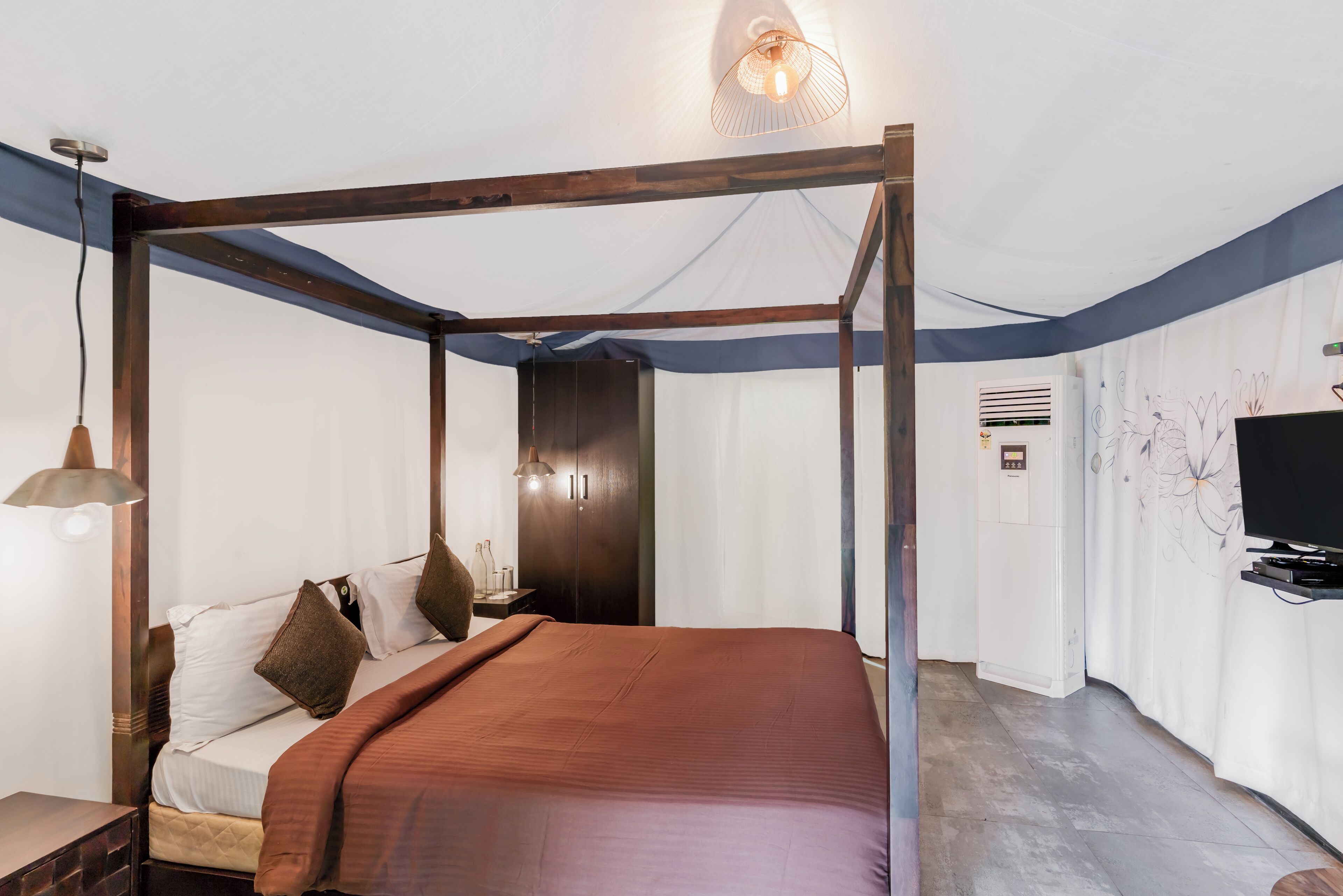 Een bed met hemelbed, met een bruin beddekleed, een nachtkastje met een lamp, een kledingkast en een televisie op een standaard.