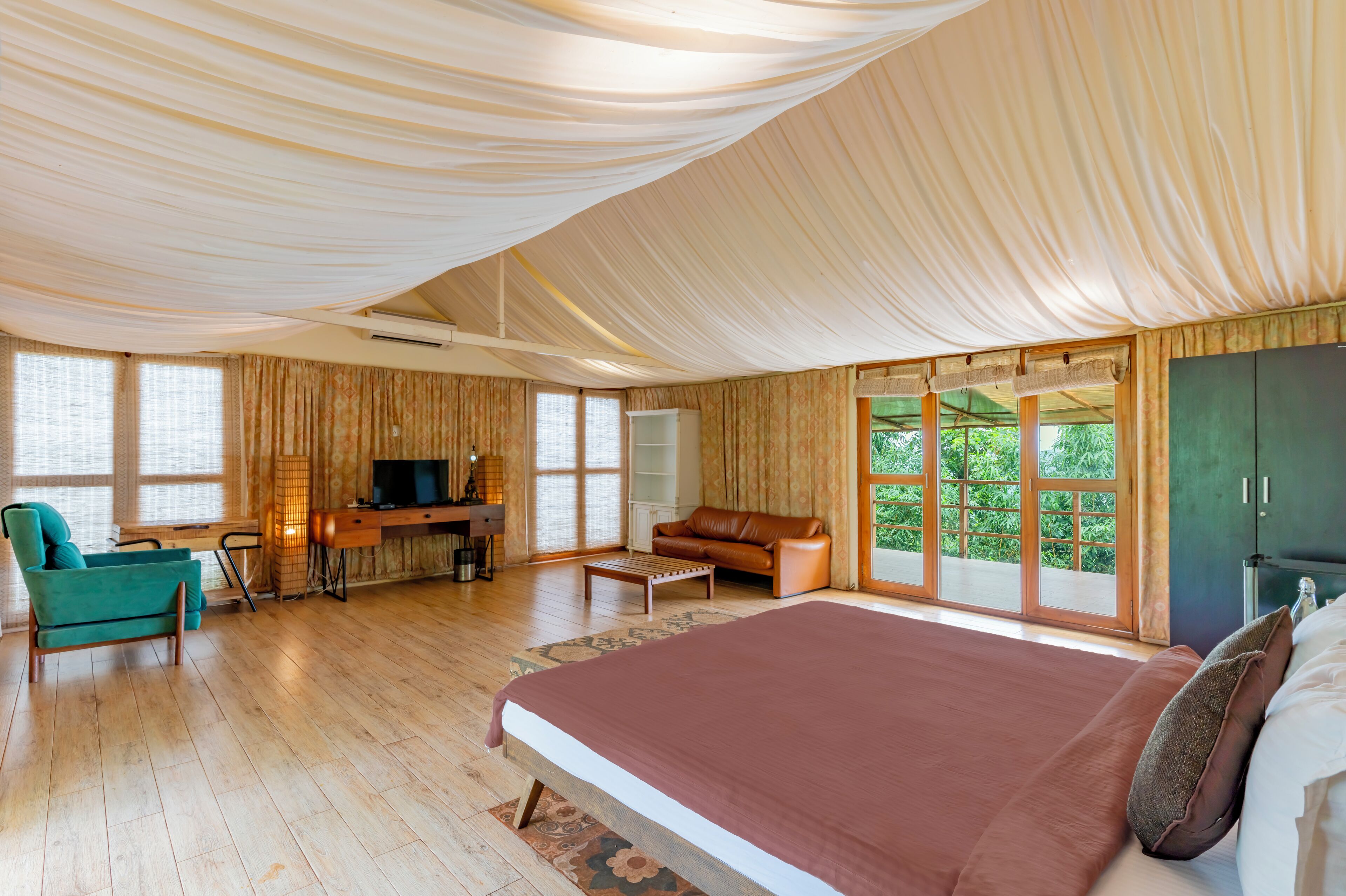 A spacious room with a large bed, a sofa, a TV, and a desk. There are large windows and a view of greenery outside.