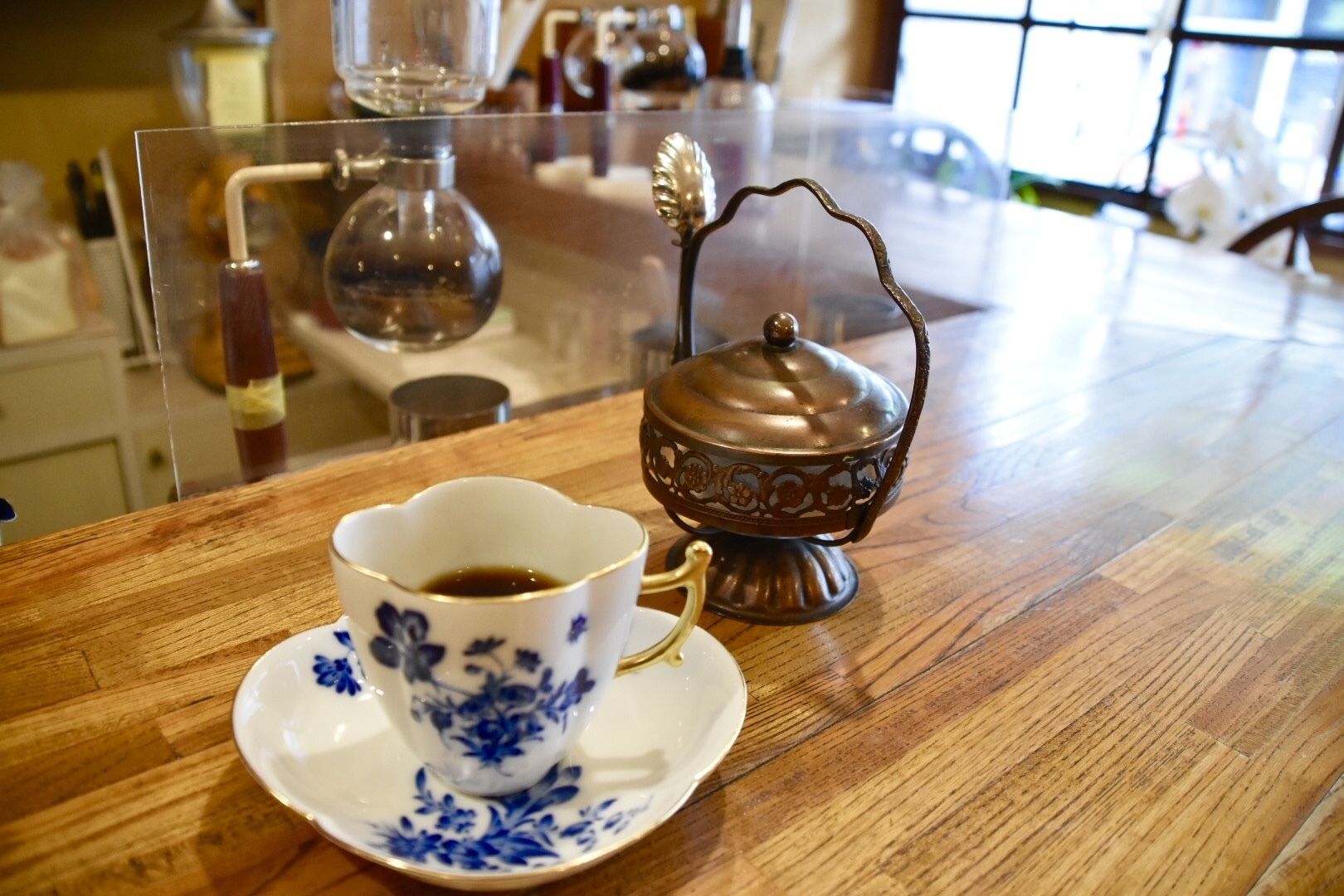 A cup of coffee on a wooden table with a decorative metal container beside it.