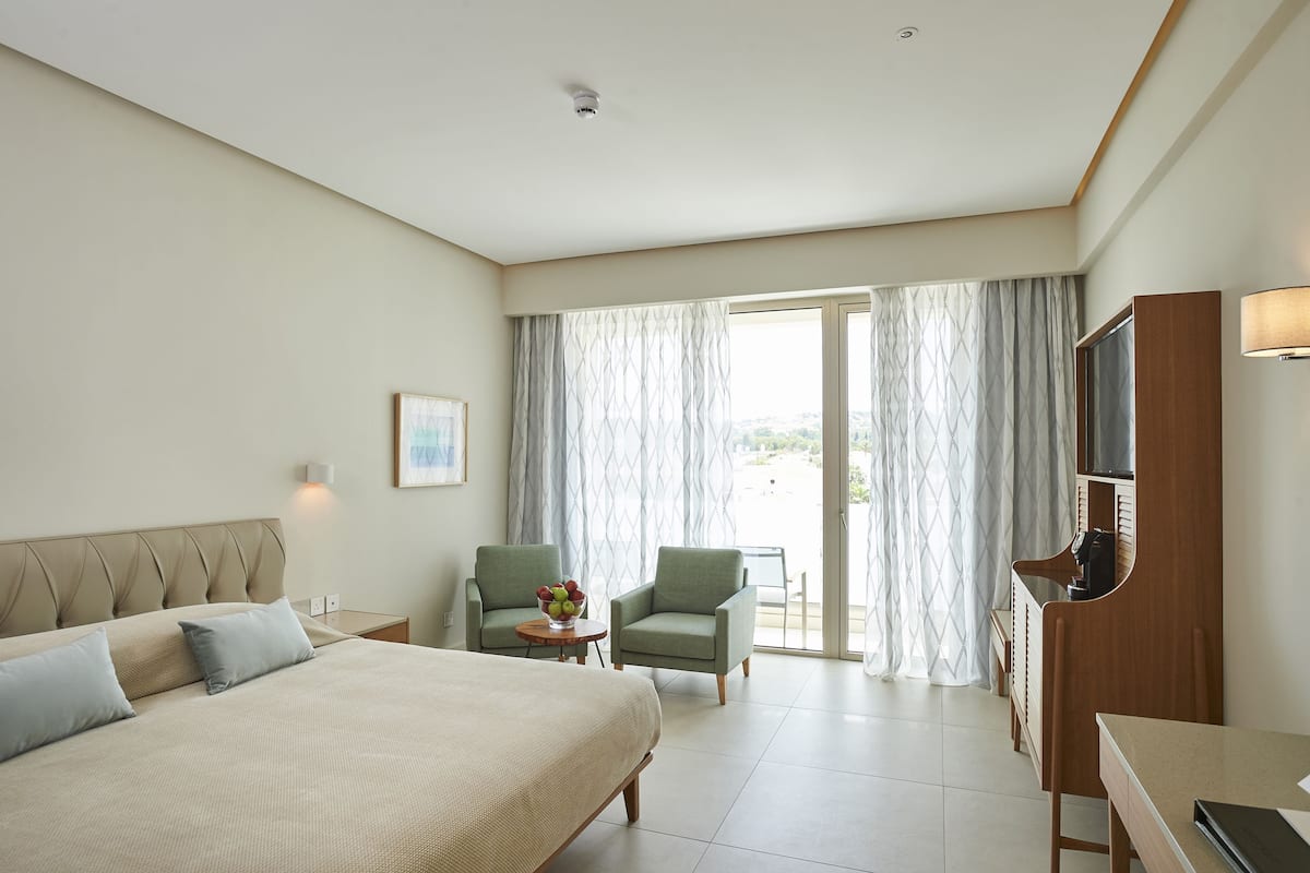 A hotel room with a bed, two armchairs, a small table with a vase, a TV, and a sliding glass door leading to a balcony.