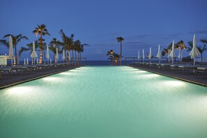 Zona de piscina con sillones de descanso y sombrillas, palmeras y vista al océano al atardecer.