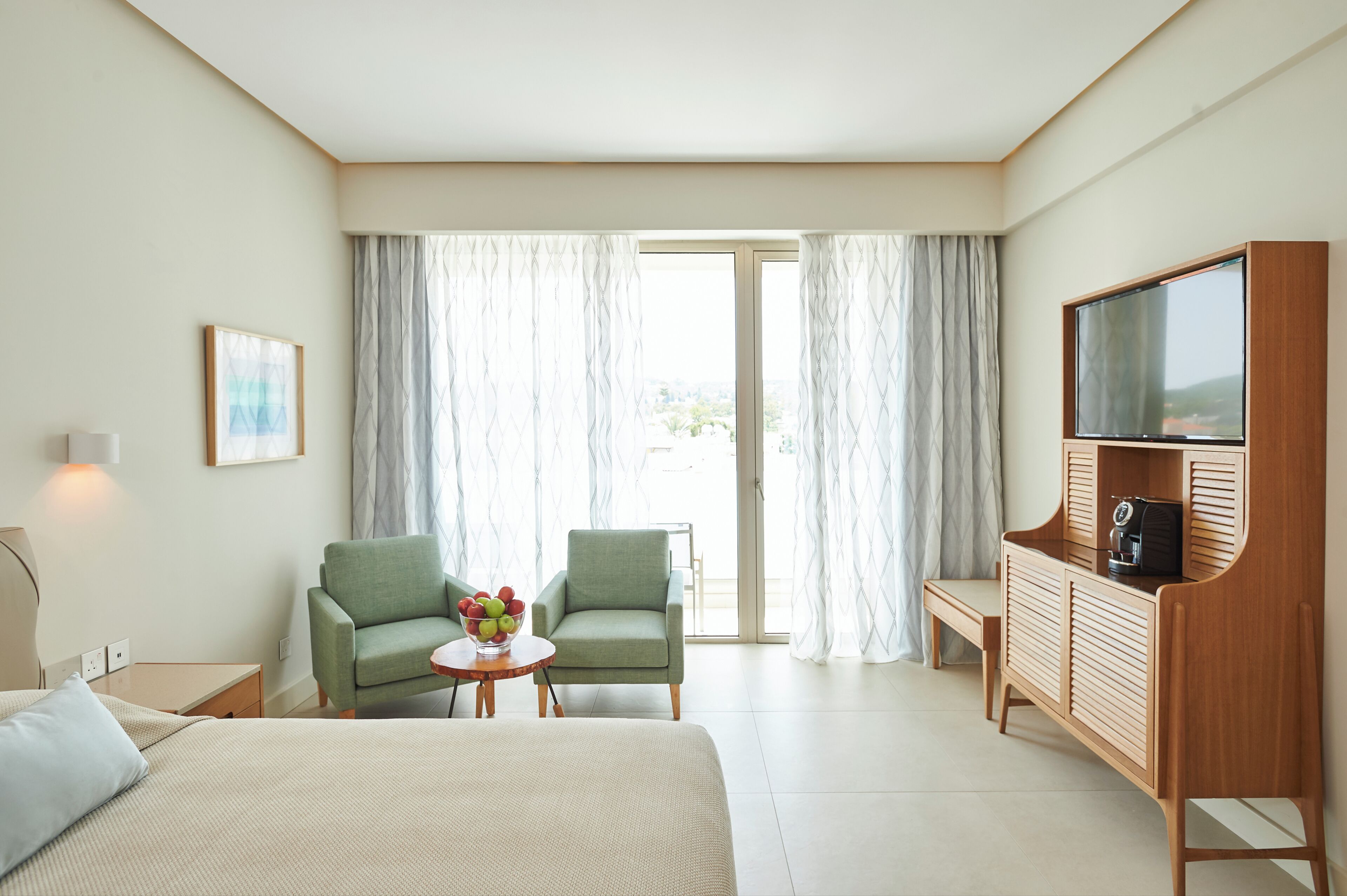 A hotel room with a bed, two chairs, a small table with fruit, a TV, and a sliding glass door to the outside.