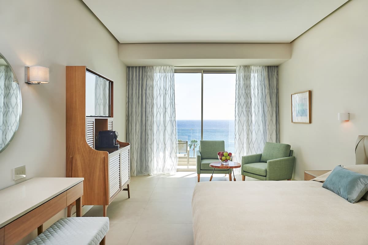 A hotel room with a bed, a desk, a chair, a small table with fruit, and a view of the sea.