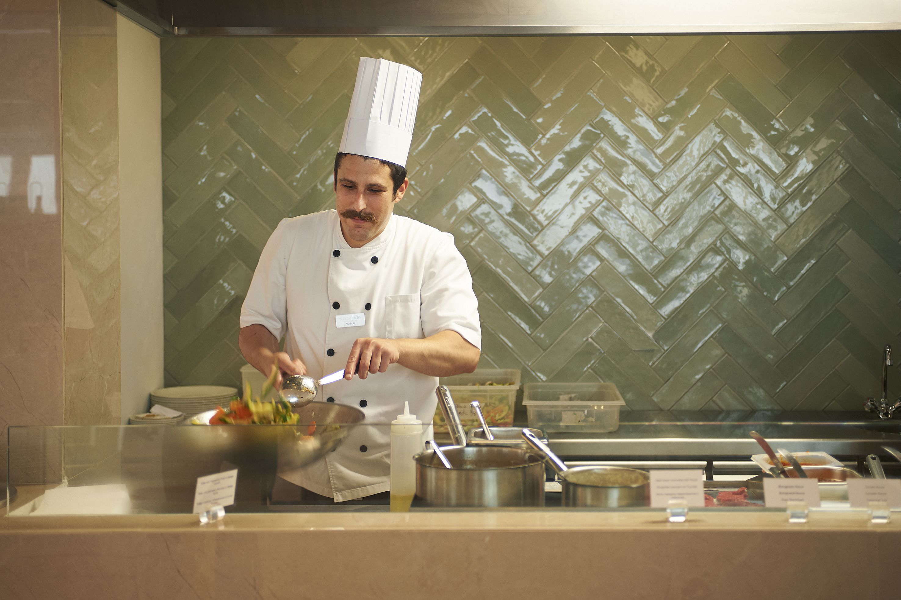 A chef in a white uniform with a tall hat, preparing food in a kitchen with a herringbone patterned backsplash.