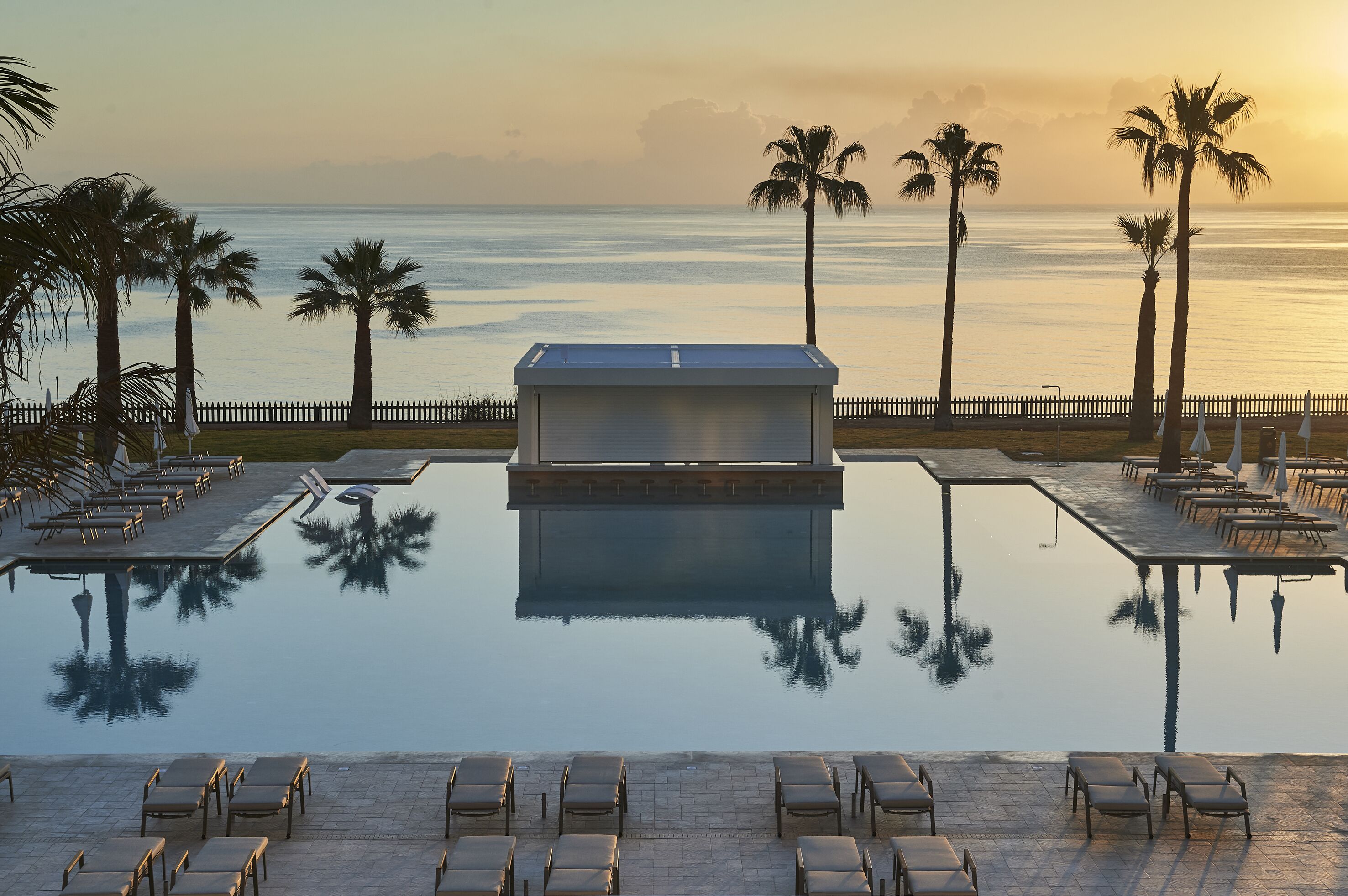 A poolside area with lounge chairs and a small building, palm trees, and a view of the ocean.