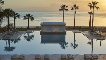 A poolside area with lounge chairs and a small building, palm trees, and a view of the ocean.