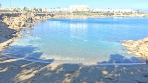 A coastal beach with clear water, rocky shoreline, and buildings in the background. - Sunrise Jade - Adults Only (Paralimni)