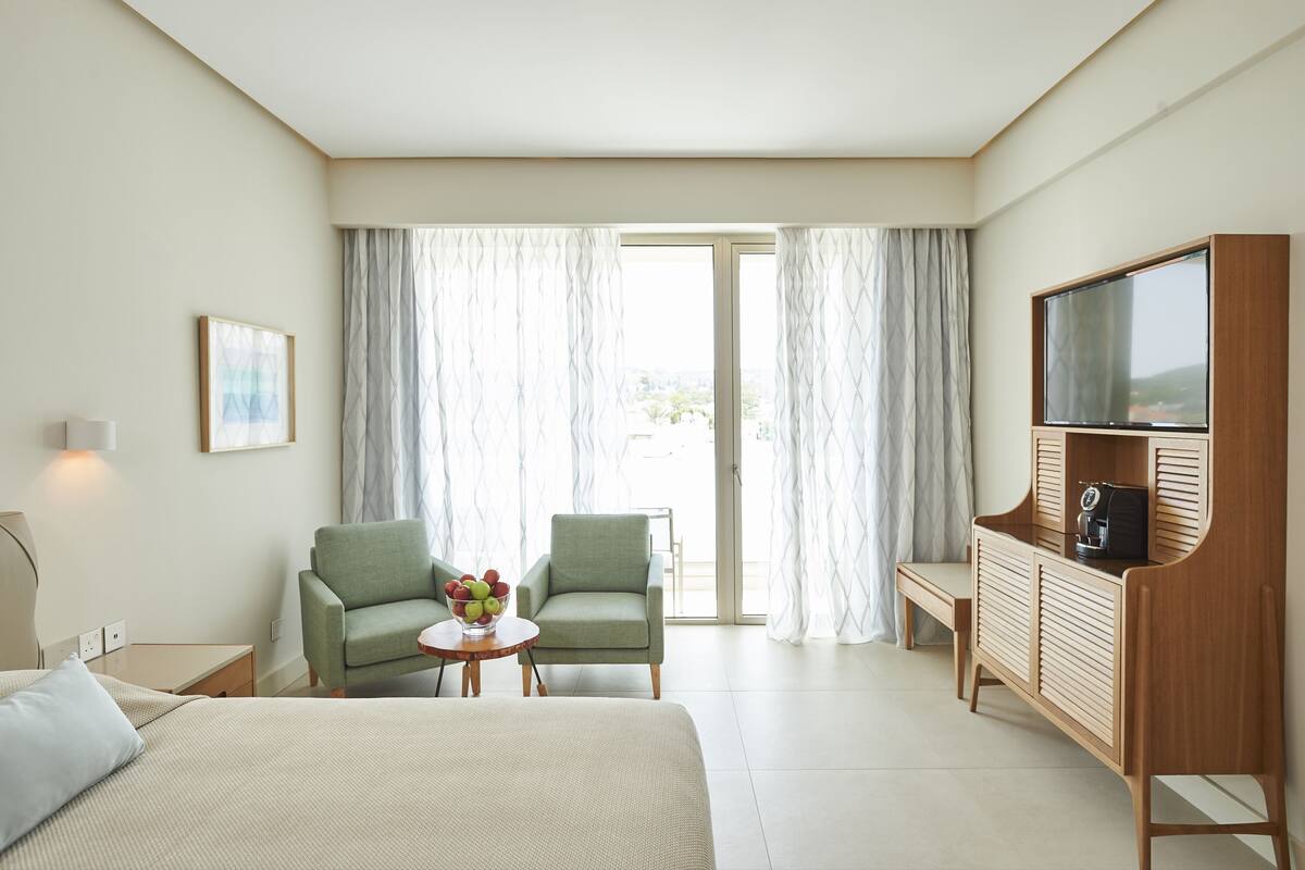 A hotel room with a bed, two chairs, a small table with fruit, a TV, and a sliding glass door.