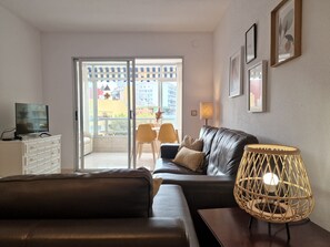 A living room with a leather sofa, a TV on a cabinet, a wicker lamp, and a view of a balcony with chairs.