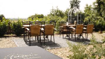 An outdoor dining area with wicker chairs and tables, surrounded by greenery and a decorative metal structure.