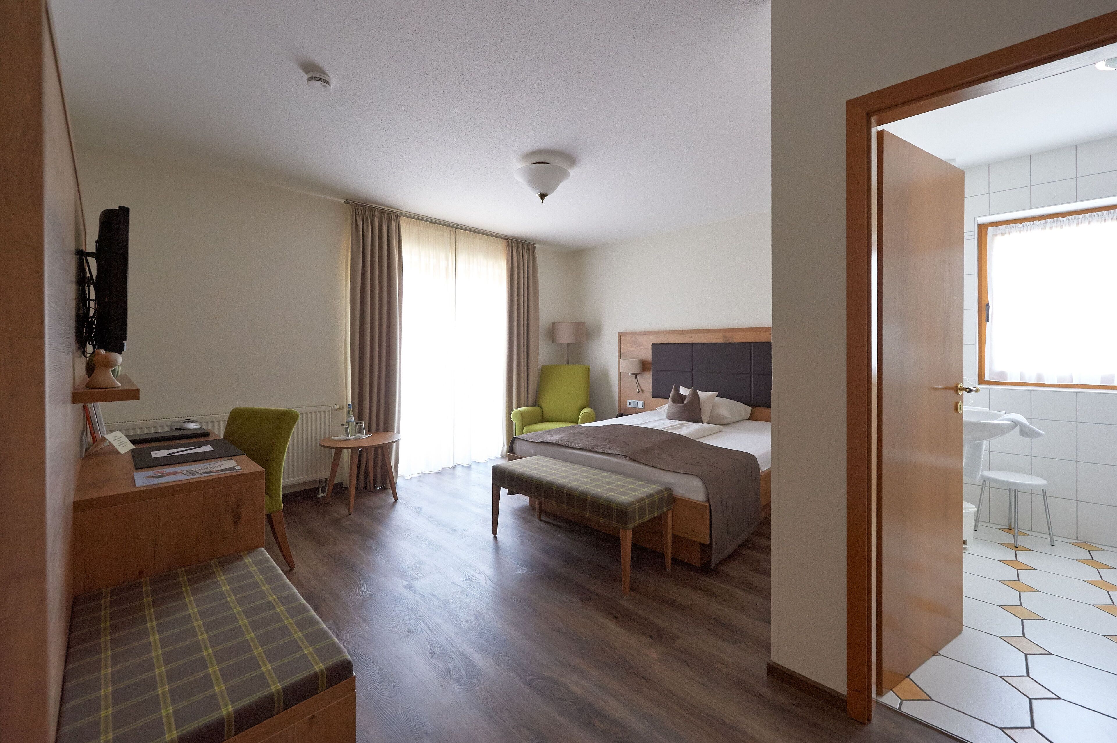 Ein Hotelzimmer mit Bett, Schreibtisch mit Stuhl, einer Bank, einem kleinen Tisch und einem Fenster mit Vorhängen.