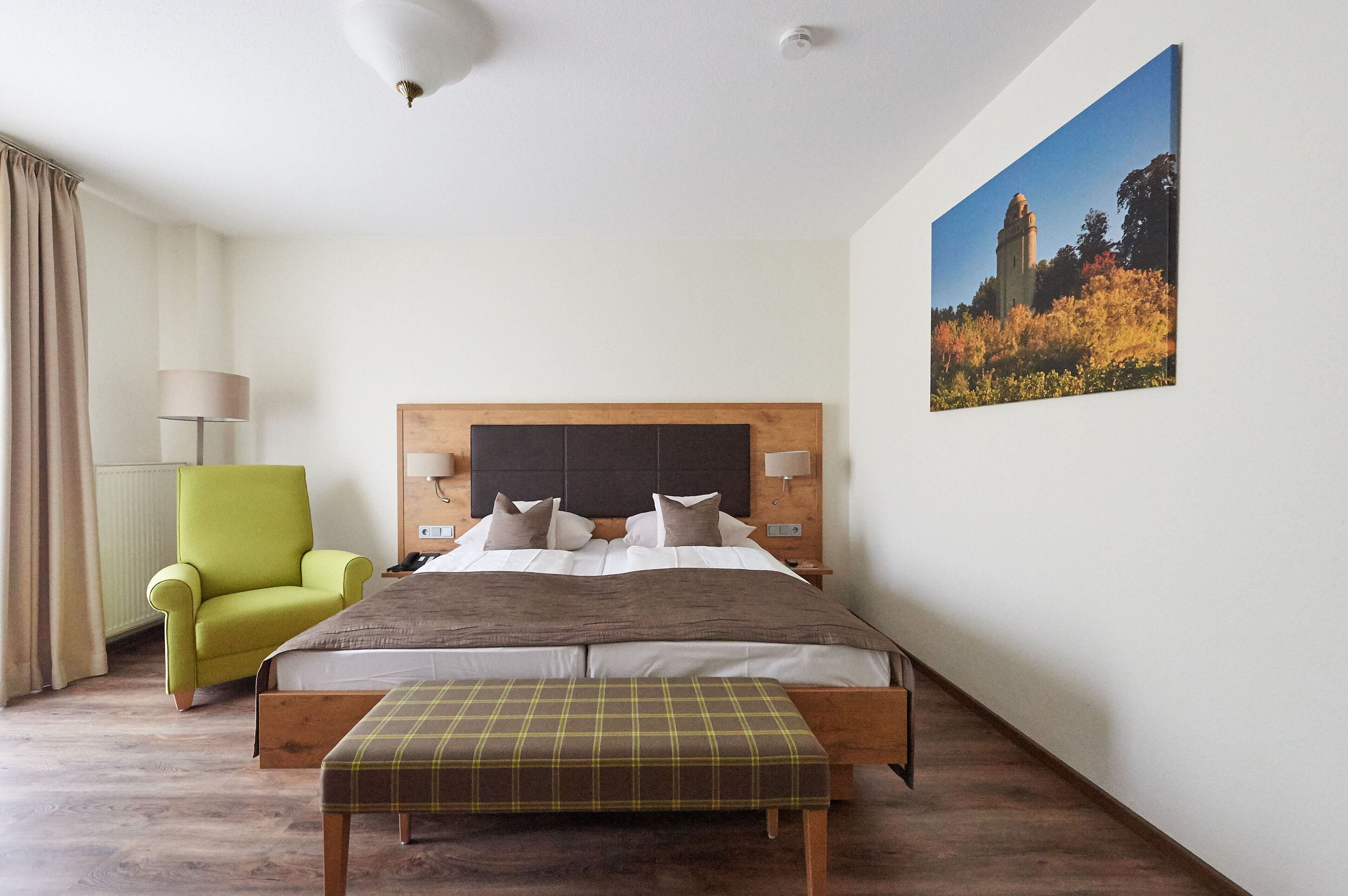A bedroom with a bed, a green armchair, a bench, a nightstand, a lamp, and a painting on the wall.