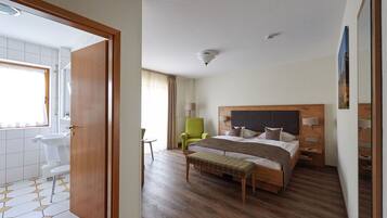 Habitación de hotel con una cama de madera, un sillón verde, una mesita de noche de madera, un lavamanos blanco y un espejo.