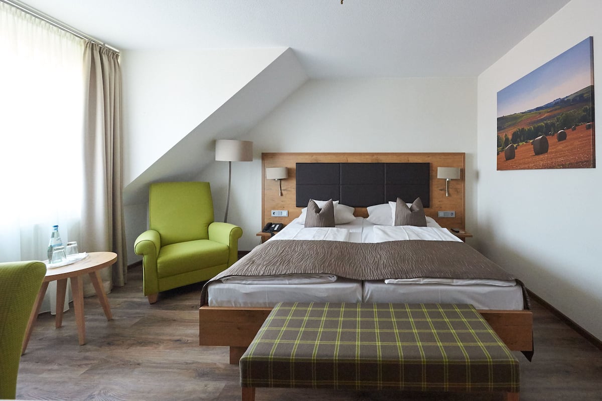 A hotel room with a bed, a green armchair, a small table with a bottle and glasses, and a painting on the wall.
