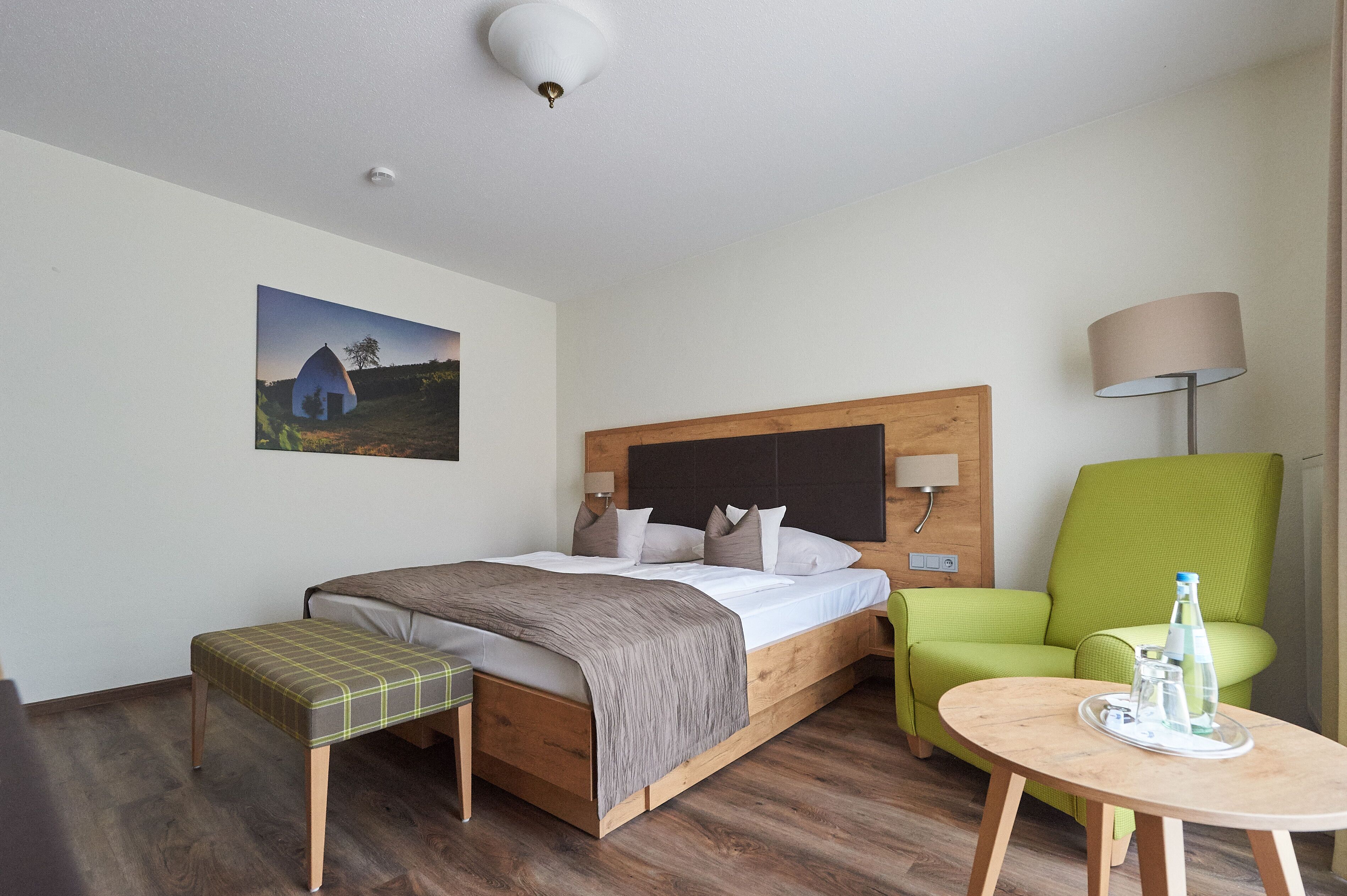Quarto de hotel com cama de madeira, uma poltrona verde, uma pequena mesa com garrafa e copos, e um quadro na parede.