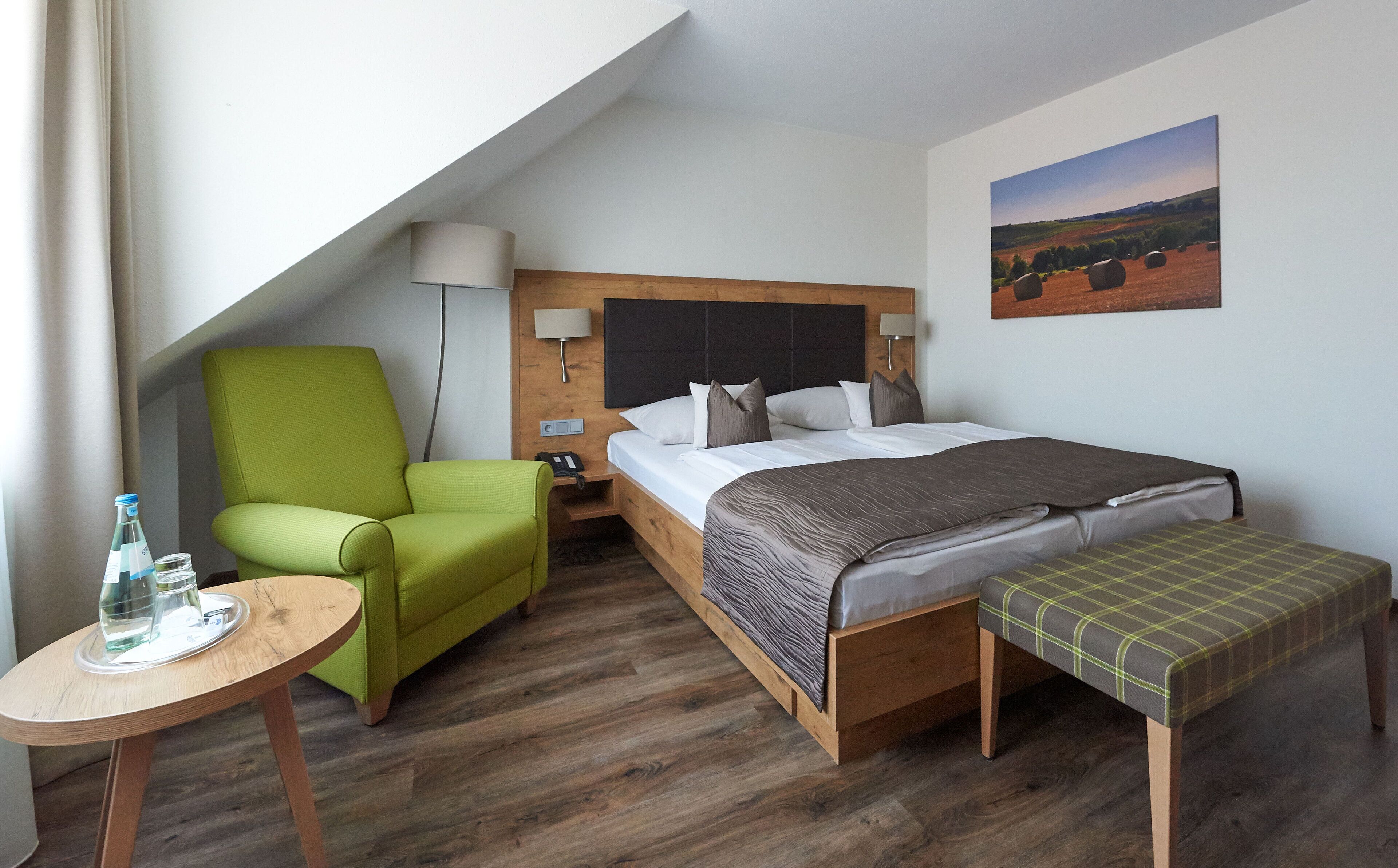 A hotel room with a bed, a green armchair, a small table with a bottle, and a painting on the wall.