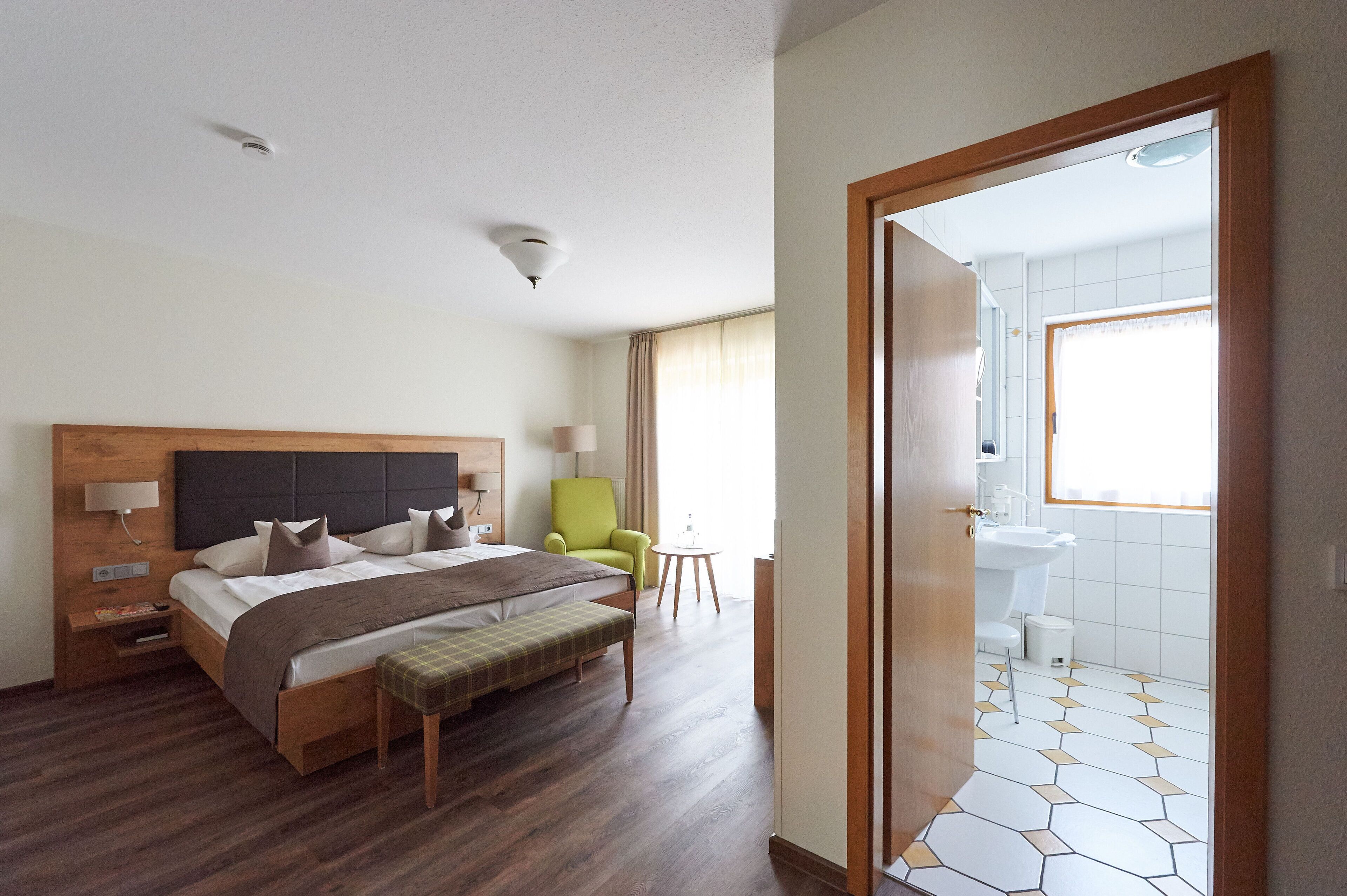 Habitación de hotel con cama, mesita de noche, lámpara, silla y vista al baño.