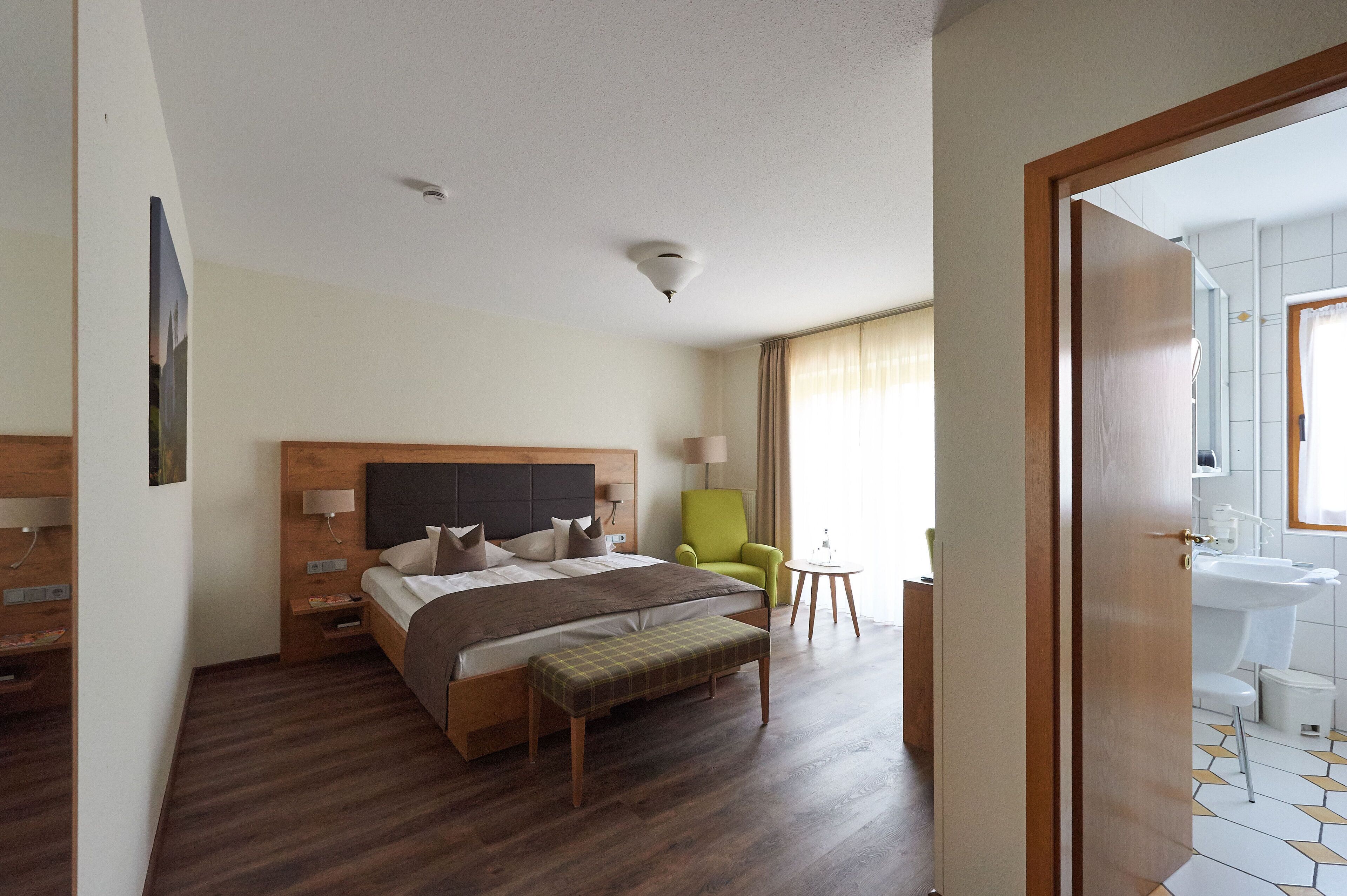 A hotel room with a bed, a bench, a chair, a bedside table, a lamp, and a view of the bathroom.