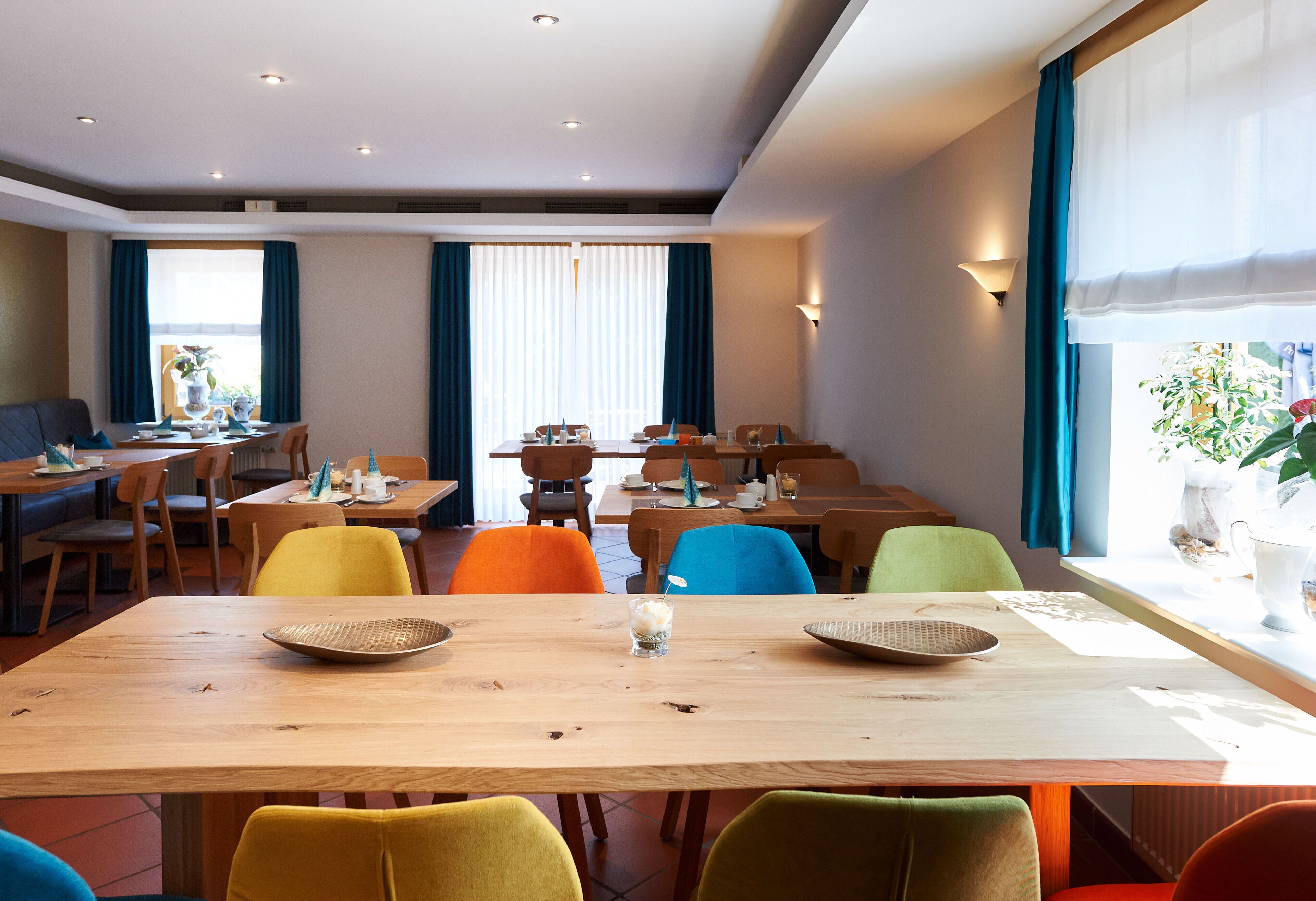 A hotel dining area with multiple tables and chairs, large windows with curtains, and a view of the outdoors.