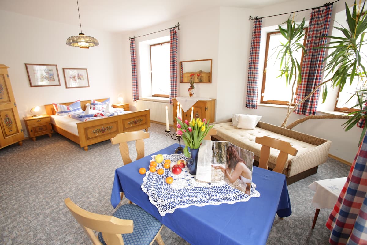 A cozy room with a bed, a sofa, a dining table with a blue tablecloth, and a window with curtains.