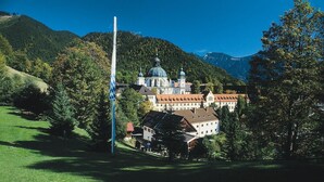 A historic building with a prominent dome and adjacent smaller structures, surrounded by lush greenery and hills. - Gästehaus Ethiko (Ettal)