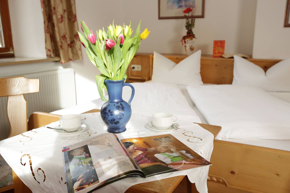 A neatly arranged hotel room with a bed, a bedside table with a vase of flowers, two cups, and a magazine on the table.