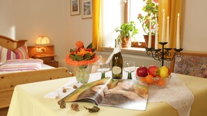 A neatly arranged bedroom with a bed, bedside table, lamp, and a table with a magazine, flowers, and fruit. - Gästehaus Ethiko (Ettal)
