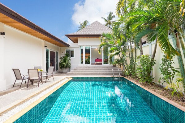 A swimming pool with a tiled edge, a covered patio area with chairs, and a building with a sloped roof and steps leading to a door. - Thai Balinese 3BR Pool Villa Alex Rawai (Rawai)