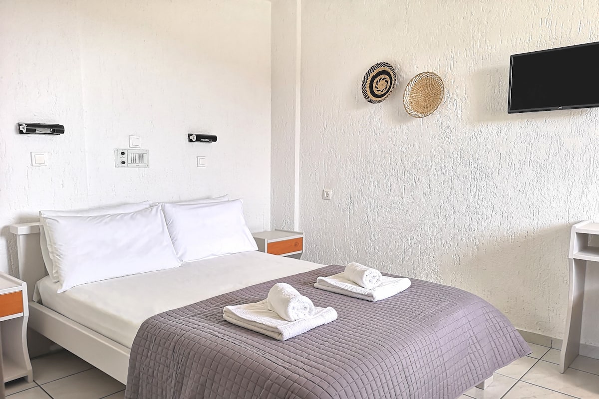 Una cama bien tendida con almohadas blancas y una colcha acolchada gris, una mesita de noche con lámpara, un televisor de pared y dos elementos decorativos colgantes.