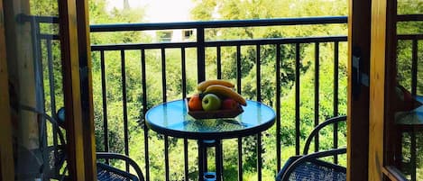 Varanda com mesa e cadeiras, uma tigela de frutas e vista para prédios e vegetação.