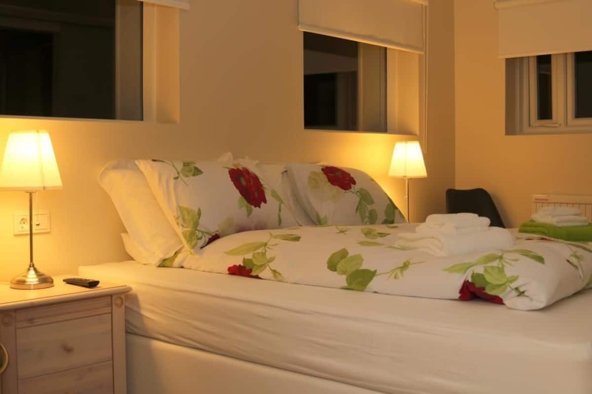 A neatly made bed with floral bedding, two bedside lamps, a nightstand with a remote control, and a mirror above the bed.