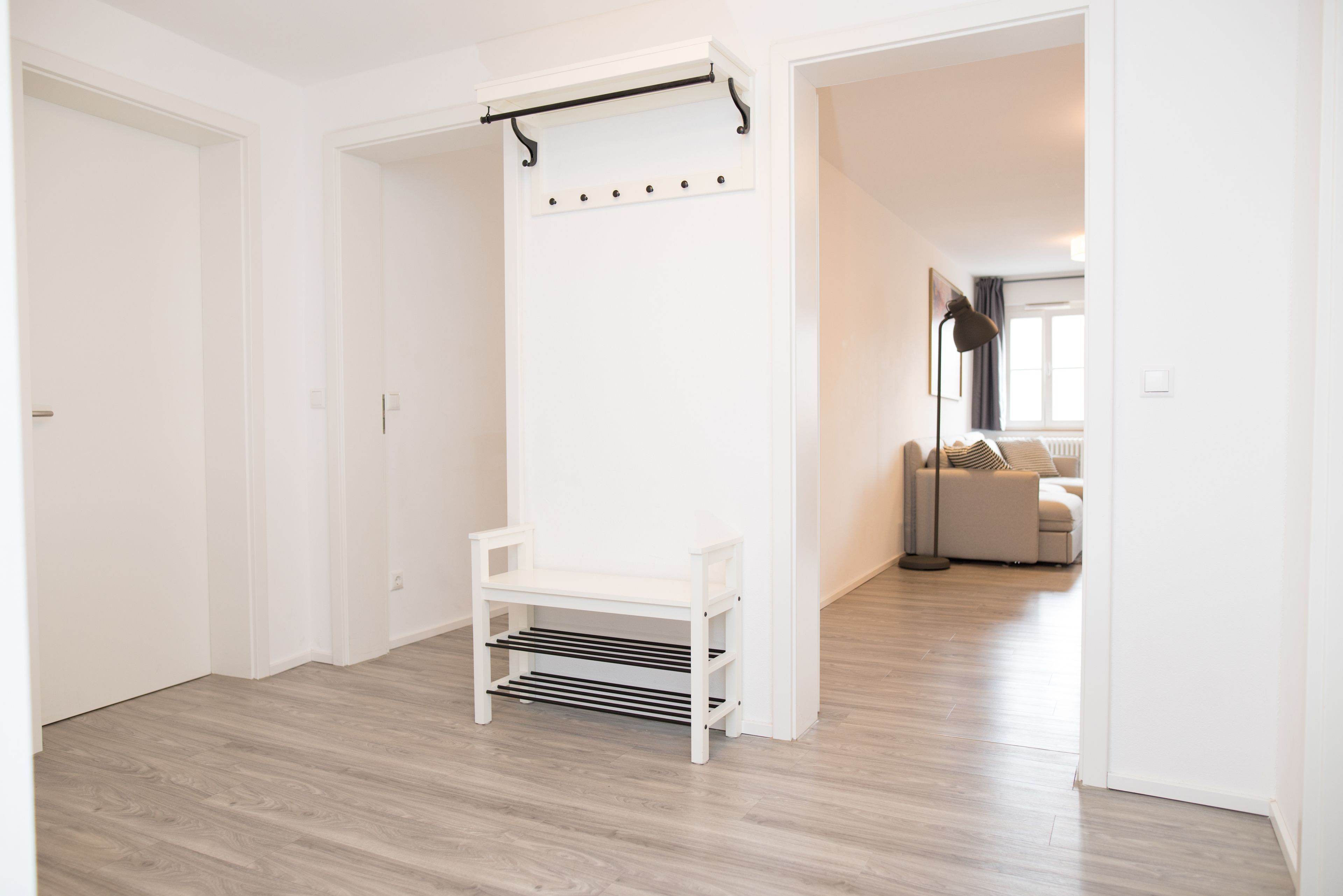 A hallway with a white bench, a coat rack, and a door leading to a living area with a sofa and a lamp.