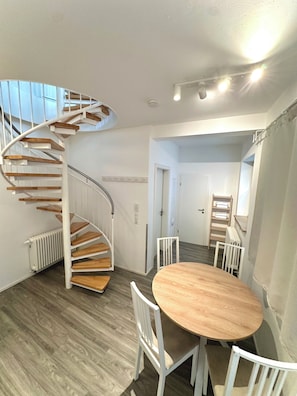 A spiral staircase with wooden steps and white railings, a round wooden table with chairs, and a view of a door leading to another room. - Arnulfsplatz Aparts (Regensburg)