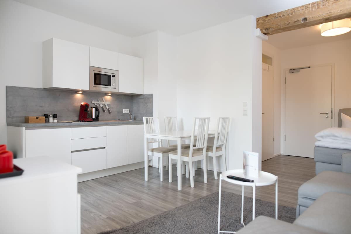 A modern kitchen with white cabinets, a microwave, and a coffee maker. There is a dining table with chairs and a small white side table.