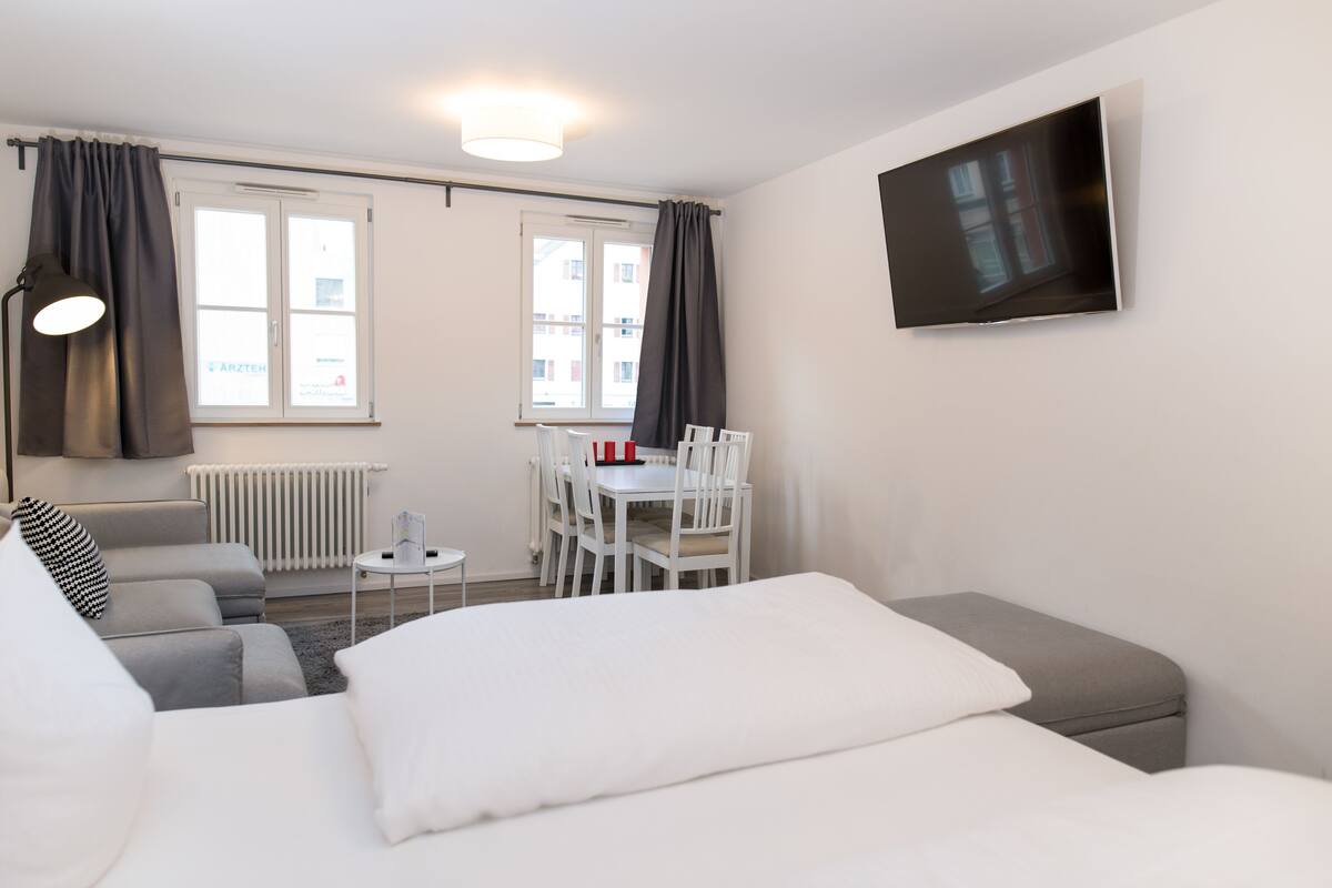 A modern hotel room with a bed, a sofa, a dining table with chairs, a TV on the wall, and a window with curtains.