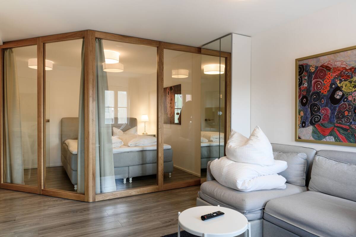 A modern hotel room with a glass-enclosed sleeping area, a grey sofa, a white round table, and a colorful abstract painting on the wall.