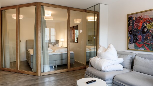 A modern hotel room with a glass-enclosed sleeping area, a grey sofa, a white round table, and a colorful abstract painting on the wall. - Arnulfsplatz Aparts (Regensburg)
