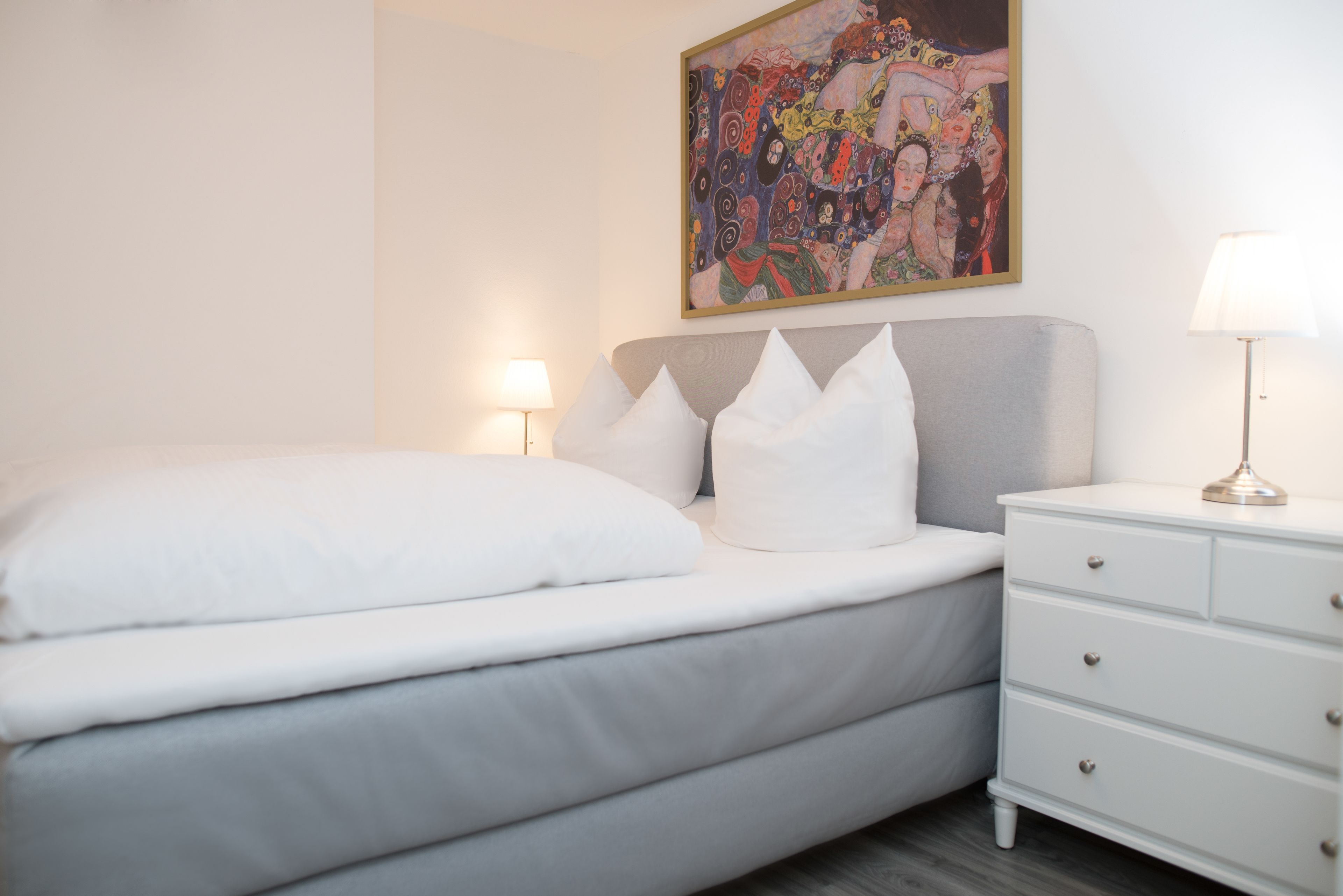 Een netjes opgemaakt bed met witte lakens en kussens, een nachtkastje met een lamp, en een kleurrijk abstract schilderij aan de muur.