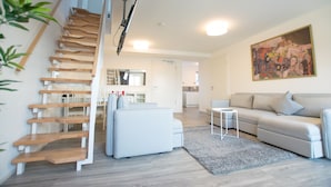 A modern living room with a staircase, a grey sofa, a small side table, and a painting on the wall. - Arnulfsplatz Aparts (Regensburg)