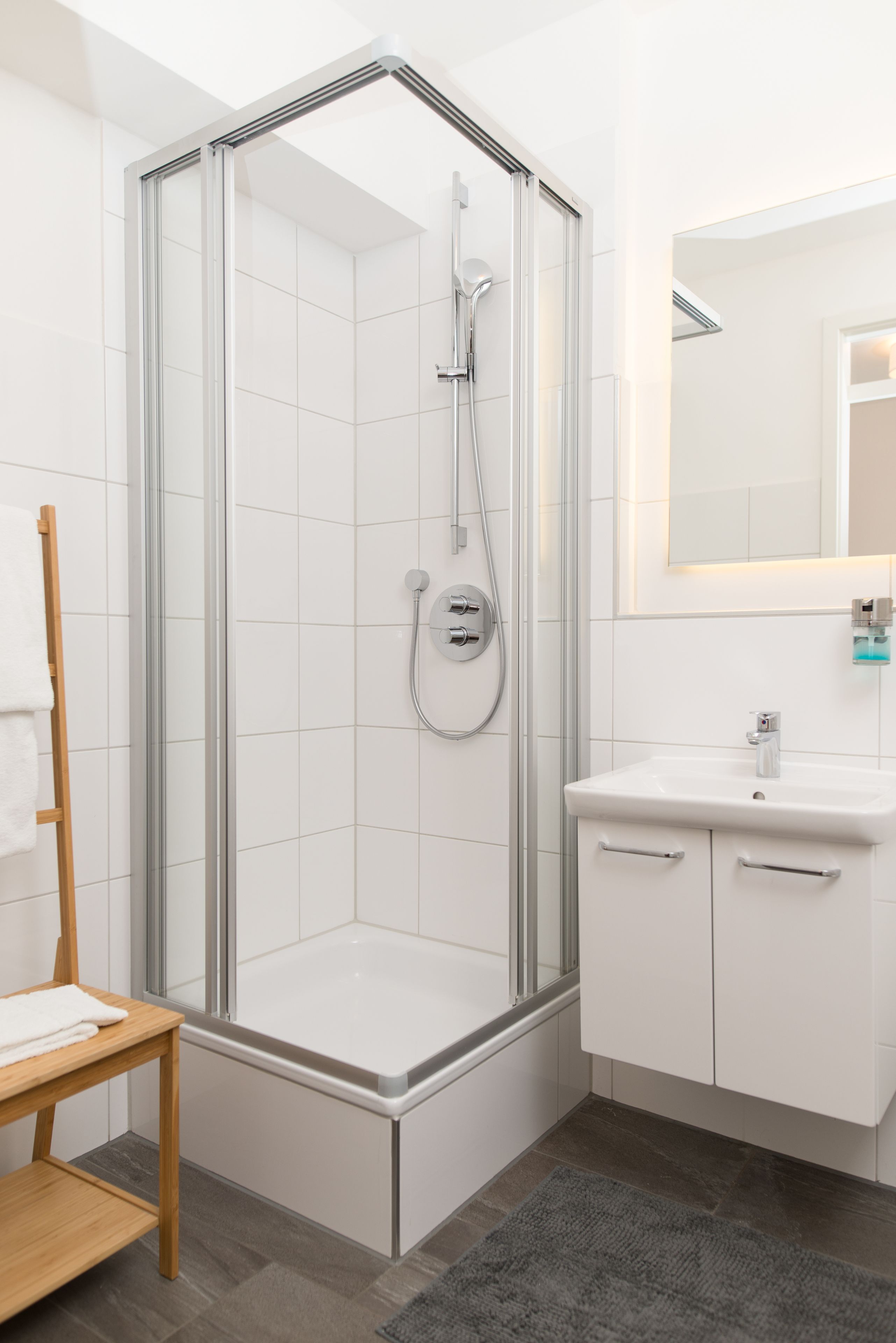 A modern bathroom with a glass-enclosed shower, a white sink, and a wooden towel rack.