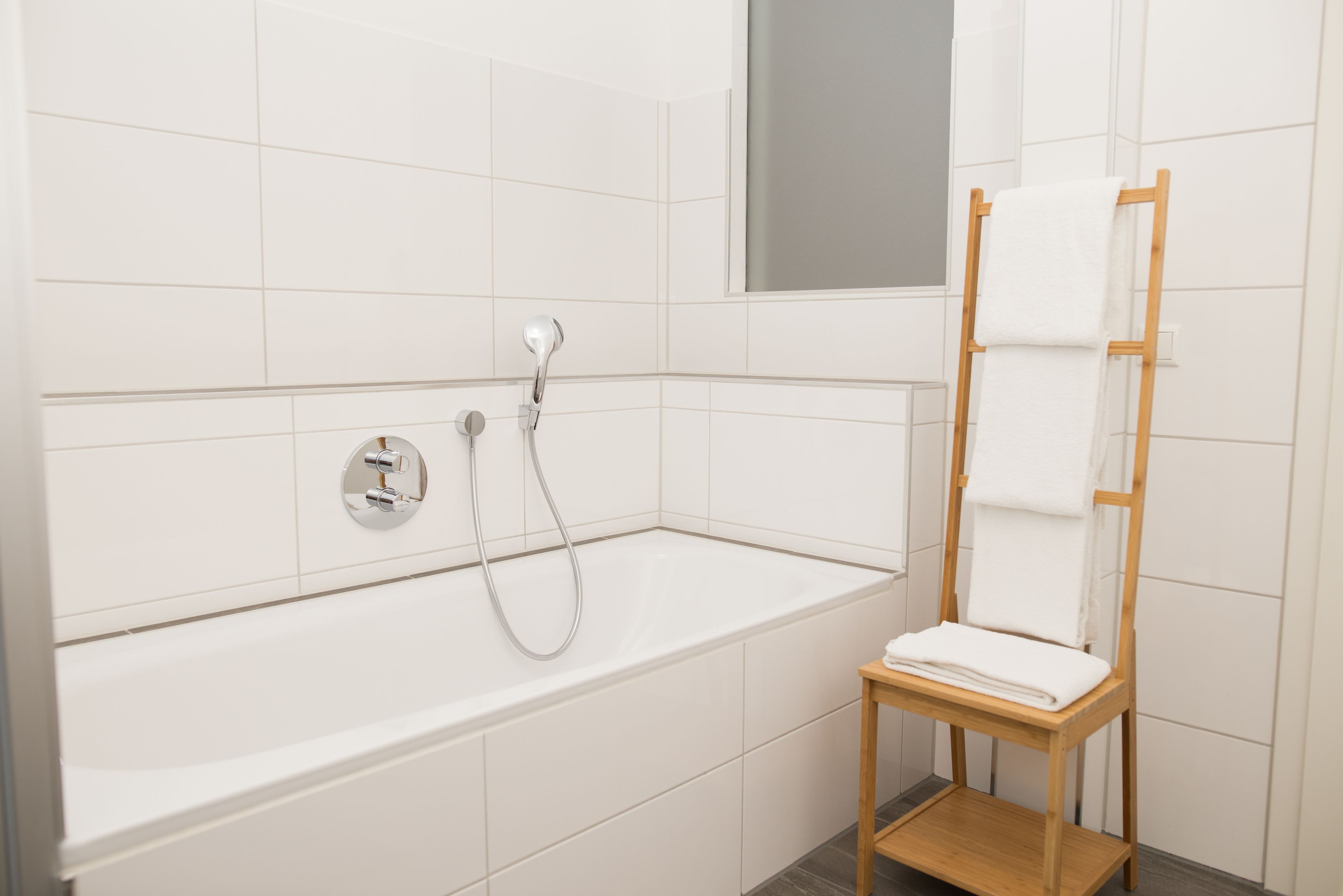 A modern bathroom with a white bathtub, a wooden towel rack, and a mirror above the tub.