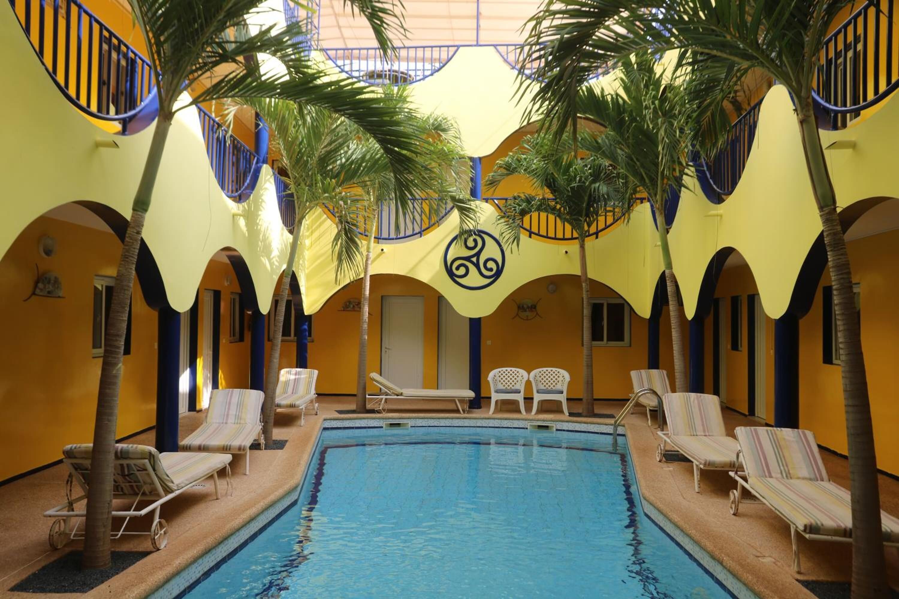 An indoor swimming pool with lounge chairs and palm trees, surrounded by yellow walls and blue railings.