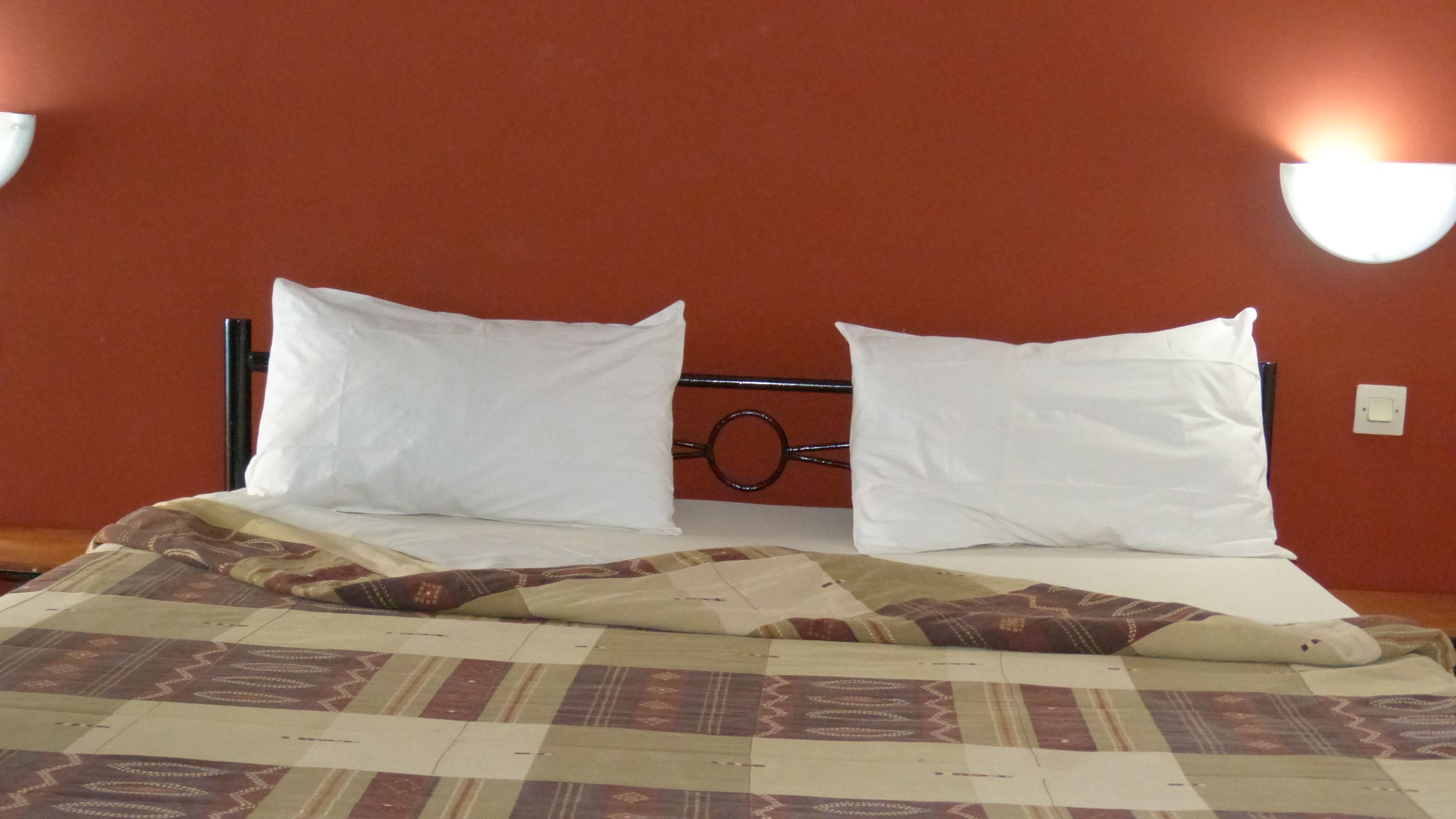 Un letto con cuscini bianchi e una coperta fantasia, una testiera in metallo e una lampada fissata al muro.