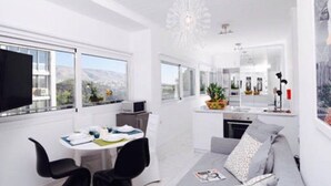A modern, well-lit living room with a grey sofa, a dining table with chairs, and a kitchen area in the background. - A Diamont with amazing view (Athens)