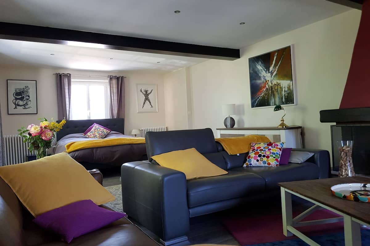 Una sala de estar moderna con un sofá, sillones y una mesa de centro. Se ve una cama con cabecera y una ventana con cortinas. Un cuadro está en la pared, y se observa un radiador.