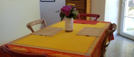 Un coin repas avec une table recouverte d’une nappe jaune, entourée de chaises en bois. Un vase rempli de fleurs est posé sur la table.