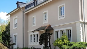 A two-story building with a beige facade, multiple windows with white frames, a small balcony, and surrounding greenery. - La Roseraie (Sentheim)