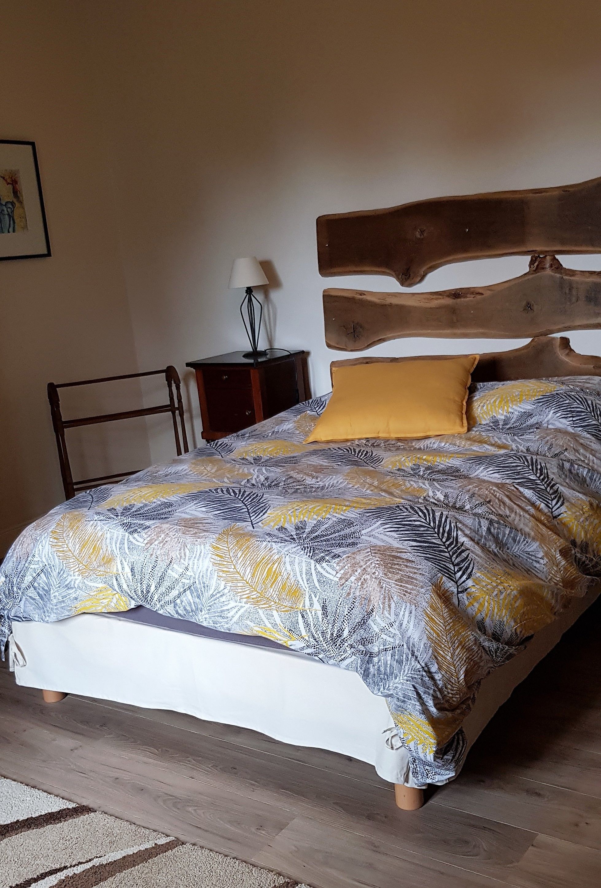 A bed with a leaf-patterned bedspread, a wooden headboard, a small bedside table with a lamp, and a chair.