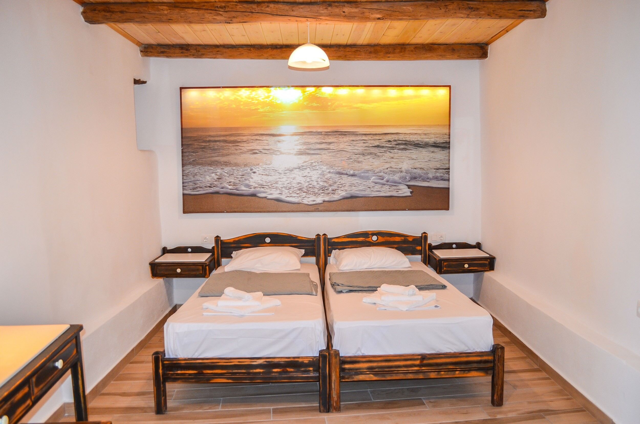 A double bed with wooden headboards and nightstands, a painting of a sunset over the sea, and a wooden ceiling.