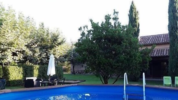 A swimming pool with a ladder, surrounded by trees and a patio area with chairs and an umbrella.